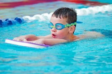 Ein Junge mit Schwimmbrille schwimmt auf einem Schwimmbrett in einem blauen Pool. Wassertropfen laufen von seinem Kopf.