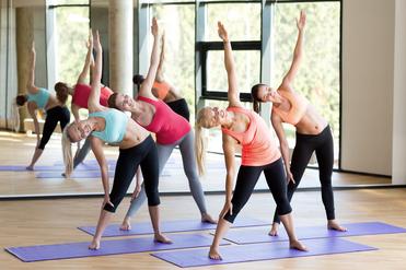 Vier Frauen in Sportkleidung machen seitliche Dehnübungen auf violetten Yogamatten in einem hellen Raum.